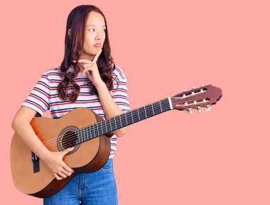 Genç güzel Çinli kız klasik gitar çalıyor. Ciddi bir yüz ifadesi var. Soru sorarken çenesi üzerine kafa karıştırıcı fikirler düşünüyor. 
