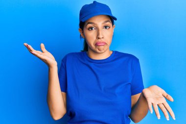 Güzel esmer kadın teslimat üniforması giyiyor. Hiçbir şeyden haberi yok ve elleri ve kolları havada kafası karışık bir ifade takınıyor. şüphe kavramı. 