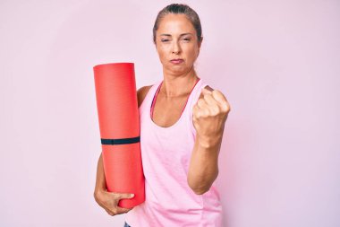 Middle age hispanic woman holding yoga mat annoyed and frustrated shouting with anger, yelling crazy with anger and hand raised 