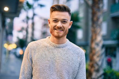 Genç beyaz adam gülümseyerek şehirde mutlu mesut duruyor..