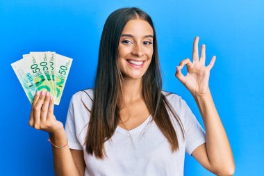 Genç İspanyol kadın İsrail şekellerini tutuyor. Parmaklarıyla imza atıyor, gülümseyerek mükemmel bir sembol oluşturuyor. 