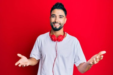 Young man with beard listening to music using headphones smiling cheerful with open arms as friendly welcome, positive and confident greetings 
