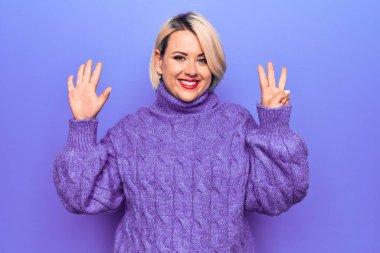 Güzel sarışın ve cüsseli bir kadın mor arka planda balıkçı yaka kazak giyiyor ve parmaklarını sekiz numarayı gösterirken kendinden emin ve mutlu bir şekilde gülümsüyor..