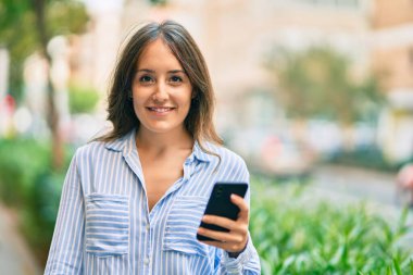 Genç İspanyol kadın şehirde akıllı telefon kullanarak mutlu bir şekilde gülümsüyor..