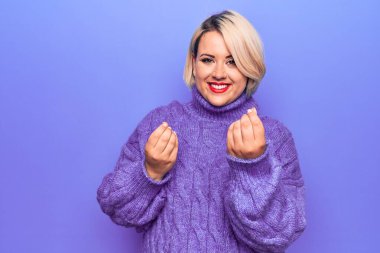 Güzel sarışın ve cüsseli bir kadın. Mor arka planda balıkçı yaka kazak giyiyor. Elleriyle para hareketi yapıyor.