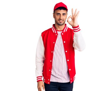 Beyzbol forması giyen genç İspanyol adam olumlu gülümsüyor ve el ve parmaklarıyla imza atıyor. başarılı ifade. 