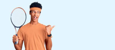 Genç Afro-Amerikalı adam tenis oynuyor raket tutuyor baş parmağını yana kaldırıyor ve ağzı açık gülümsüyor. 