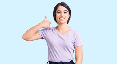 Esmer, sıradan kıyafetler giyen, gülümseyen, el ve parmaklarıyla telefon hareketi yapan, telefonda konuşan bir kız. iletişim kavramları. 