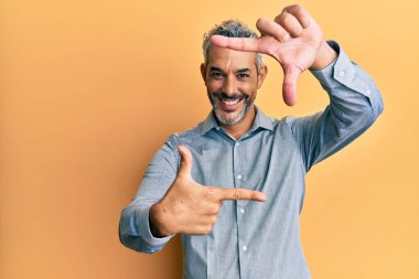 Orta yaşlı, gri saçlı, günlük kıyafetler giyen, gülümseyen, elleri ve parmakları mutlu yüzlü bir adam. yaratıcılık ve fotoğrafçılık kavramı. 