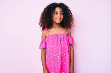 Kıvırcık saçlı, sıradan bir elbise giyen Afro-Amerikan çocuğu gülümseyerek yan tarafa bakıyor ve düşünüyor.. 