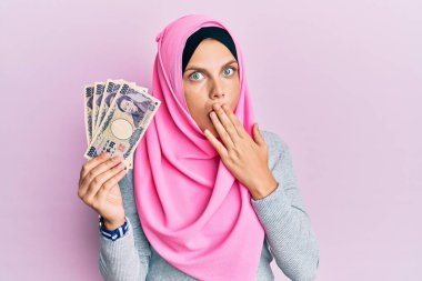 Kafkasyalı genç bir kadın, elinde Japon yen banknotları tutan, ağzı elle kaplayan, şok olmuş ve hata yapmaktan korkan, İslami başörtüsü takıyor. şaşırmış ifade 
