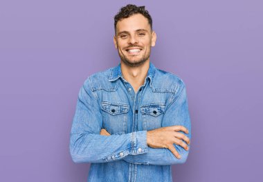 Sıradan kot ceket giyen genç İspanyol adam kollarını kavuşturup kameraya bakarken gülümsüyor. pozitif kişi. 