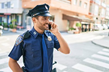 Genç, yakışıklı İspanyol polis memuru polis üniforması giyip mutlu gülümsüyor. Yüzünde tebessümle dikilip şehrin caddesinde akıllı telefondan konuşuyor..