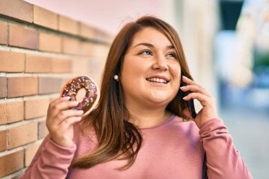 Genç Latin asıllı, iri yarı bir kadın akıllı telefondan konuşuyor ve şehirde çörek yiyor..