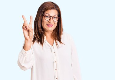 Orta yaşlı İspanyol kadın gündelik kıyafetler giyiyor. Gülümsüyor. Arkadaşça gülümsüyor ve iki numarayı işaret ediyor.