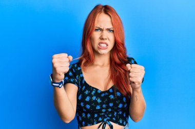 Genç, güzel, kızıl saçlı bir kadın mavi arka plan üzerine gündelik kıyafetler giymiş kızgın ve kızgın yumruklar sallıyor öfkeyle bağırırken sinirli ve öfkeli. öfke ve agresif konsept. 