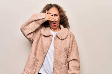 Orta yaşlı, sıradan bir ceket giyen güzel bir kadın izole edilmiş beyaz bir arka plan üzerinde duruyor. Şok içinde yüzünü ve gözlerini eliyle kapatıyor, parmaklarından korkuyla bakıyor.