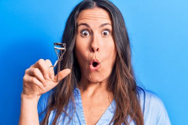 Genç, güzel esmer kadın kirpik bigudisini soyutlanmış mavi arka planda tutuyor. Şaşırmak için ağzı açık kalmış, korkmuş ve hayrete düşmüş.