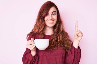 Elinde kahveyle gülümseyen genç ve güzel bir kadın bir fikir ya da soruyla mutlu bir yüzle işaret ediyor. 