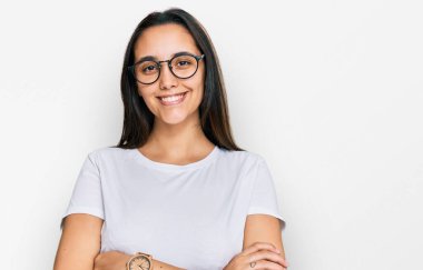 Gündelik beyaz tişört giyen genç İspanyol kadın kollarını kavuşturup kameraya bakarken gülümsüyor. pozitif kişi. 