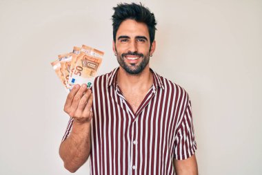 Avro banknotları tutan yakışıklı İspanyol adam pozitif ve mutlu görünüyor ve kendine güvenen bir gülümsemeyle dişlerini gösteriyor. 