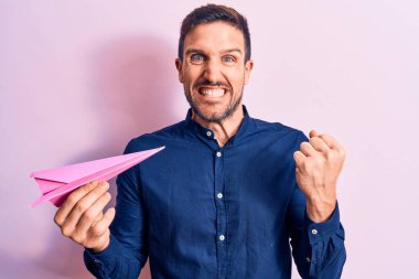 Arkaplanda pembe kağıt uçak tutan genç yakışıklı adam gururla haykırıyor, zaferi ve başarıyı kutluyor.