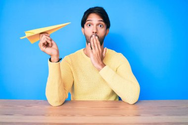 Eli kağıt uçak tutan yakışıklı İspanyol adam ağzını eliyle kapatıyor, şokta ve hata yapmaktan korkuyor. şaşırmış ifade 