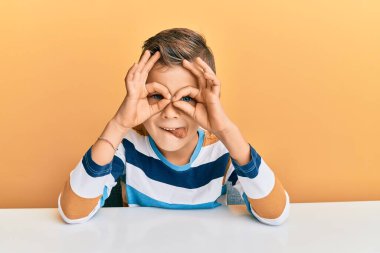 Kafkasyalı sevimli bir çocuk, günlük giysiler giymiş masanın üzerinde oturmuş dürbün gibi dil çıkartıyor, gözler parmaklarından bakıyor. çılgın ifade. 