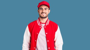 Beyzbol ceketi ve şapkası giyen yakışıklı genç adam pozitif ve mutlu görünüyor. Duruyor ve gülümsüyor. Kendine güvenen bir gülümsemeyle dişlerini gösteriyor. 
