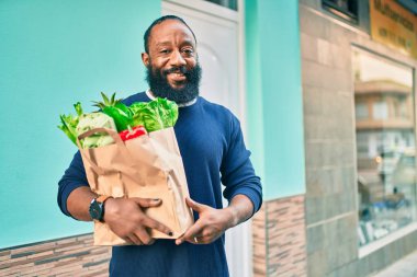 Sakallı Afrikalı Amerikalı bir adam elinde süpermarketten alınmış kağıt poşetler tutuyor.