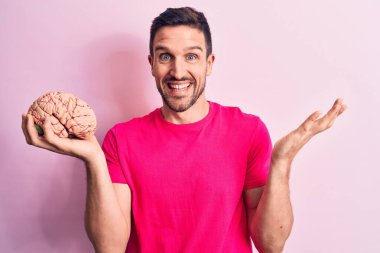 Genç yakışıklı adam beyni soyutlanmış pembe arka planda tutan mutlu bir gülümseme ve kaldırılmış bir el ile kazanan bir ifadeyle başarıyı kutlayan özenli bir hatıra istiyor.