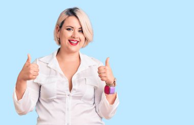 Genç sarışın ve cüsseli bir kadın, günlük tişört giyer, başarı işareti yapar, elleriyle olumlu jest yapar, gülümser ve mutlu olur. Neşeli ifade ve kazanan jest. 