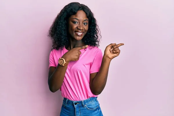 美しいアフリカの若い女性を着てカジュアルなピンクのTシャツ笑顔とカメラを見て2本の手と指で側面を指して — ストック写真