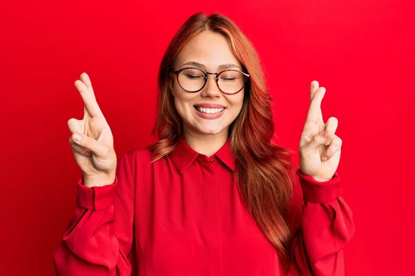 年轻美丽的红头发女人 身穿休闲服 戴着眼镜 戴在红色背景上 摆出手势 微笑着 闭上了眼睛 幸运与迷信的概念 — 图库照片