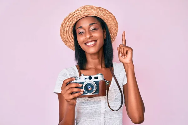 Young african american woman wearing summer hat holding vintage camera pointing finger up with successful idea. exited and happy. number one.