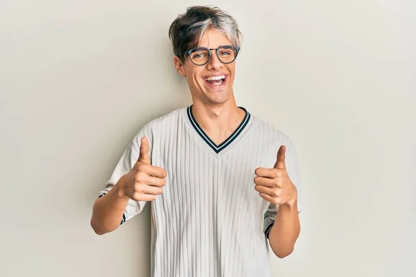 カジュアルな服と眼鏡の成功のサインを身に着けている若いヒスパニック系の男は 手で積極的なジェスチャーを行うと 親指を笑顔と幸せ 陽気な表情と勝者のジェスチャー — ストック写真
