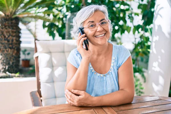 Middle age woman with grey hair smiling happy relaxing sitting at the terrace at home speaking on the phone