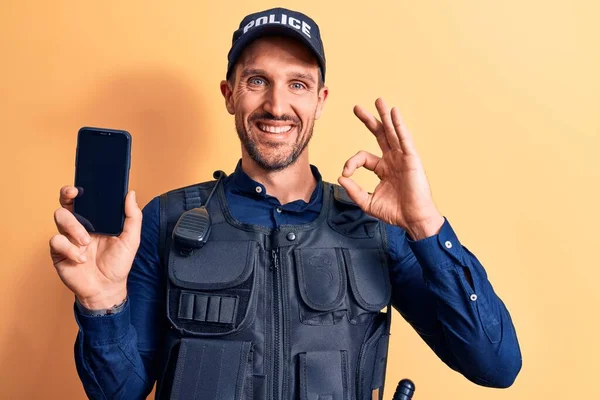 Handsome policeman wearing uniform and bulletprof holding smartphone showing screen doing ok sign with fingers, smiling friendly gesturing excellent symbol