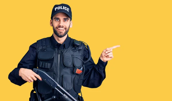 Young handsome man with beard wearing police uniform holding shotgun smiling happy pointing with hand and finger to the side