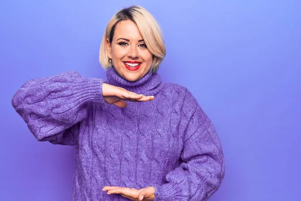 Beautiful blonde plus size woman wearing casual turtleneck sweater over purple background gesturing with hands showing big and large size sign, measure symbol. Smiling looking at the camera. Measuring concept.