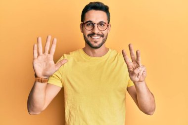 Gündelik kıyafetler giyen ve gözlük takan İspanyol genç adam sekiz numaralı parmaklarını gösterip işaret ederken kendinden emin ve mutlu gülümsüyor.. 