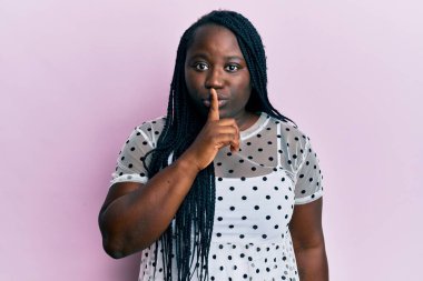 Saçları örgülü, sıradan kıyafetler giyen genç siyahi bir kadın dudakları parmaklarıyla sessiz olmak istiyor. sessizlik ve gizli konsept. 