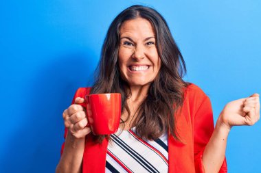 Genç ve güzel esmer iş kadını izole edilmiş mavi arka planda bir kupa kahve içiyor gururla bağırıyor, zaferi ve başarıyı kutluyor.