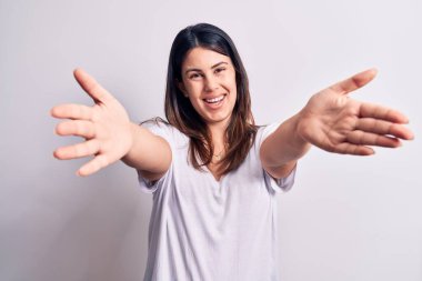 Genç, güzel, esmer, günlük tişörtler giyen bir kadın izole edilmiş beyaz arka planda dikilip kameraya bakarak sarılmak için kollarını açmış gülümsüyor. Mutluluğu kucaklayan neşeli bir ifade..