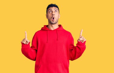 Gündelik giysiler giyen genç İspanyol adam şaşırmış ve şaşırmış bir şekilde yukarı bakıyor ve parmaklarıyla işaret ediyor ve kollarını kaldırıyor.. 