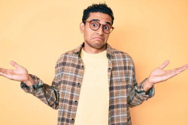 Sıradan kıyafetler giyen, gözlüklü, kafası karışık, elleri ve kolları havada genç, yakışıklı İspanyol bir adam. şüphe kavramı. 