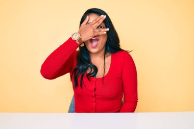 Uzun saçlı, güzel bir Latin kadın. Günlük giysiler giymiş. Masada oturuyor. Şok olmuş bir şekilde yüzünü ve gözlerini eliyle kapatıyor. 