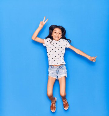 Sevimli İspanyol kız günlük kıyafetler giyip mutlu gülümsüyor. Yüzünde gülümsemeyle zıplıyor, mavi arka planda zafer işareti yapıyor.