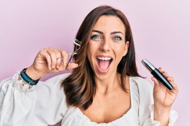Young brunette woman holding eyelash curler celebrating crazy and amazed for success with open eyes screaming excited.  clipart