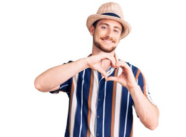 Yazlık şapka takan yakışıklı, beyaz bir adam aşka gülümseyip elleriyle kalp sembolü çiziyor. romantik kavram. 